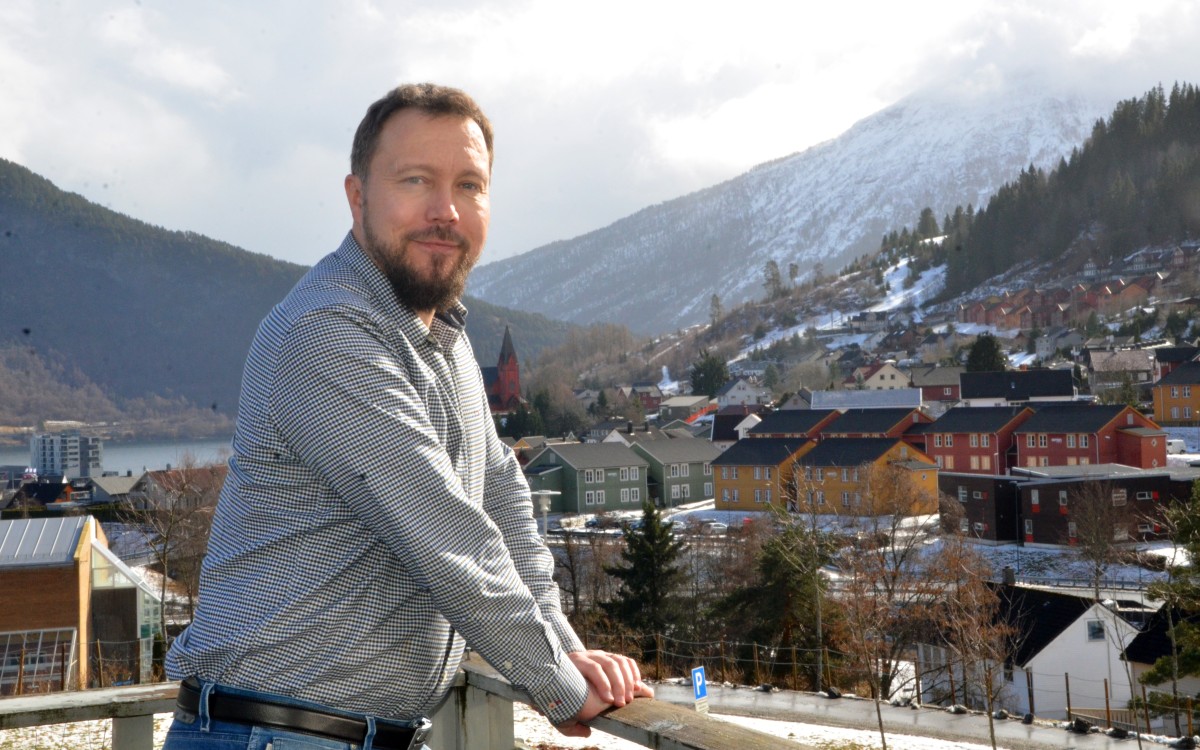 Ung mann på veranda med fjell og bygd bak seg, ser i kamera