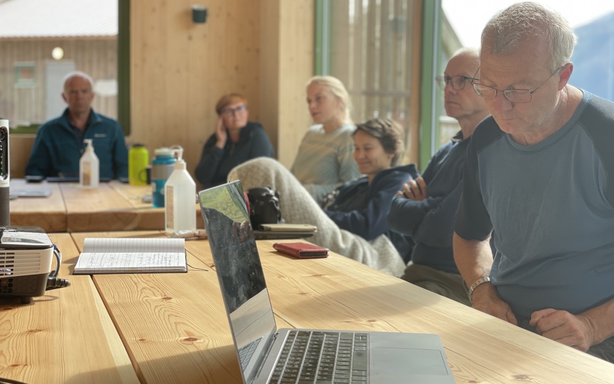 Trebord i lyst lokale. Kollegaer i møte sit og ser smilande framfor seg. Laptop i framgrunnen.