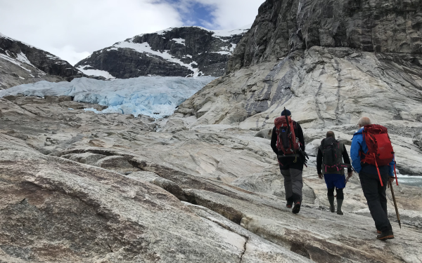 bratte svaberg framfor brefall, tre fjellkledde folk går oppover mot isen i det fjerne 