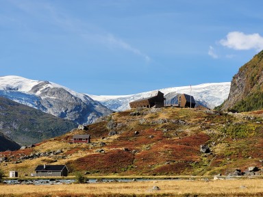 moderne hyttebygg på haug med fjell og bre bak, blå himmel over