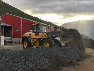 Hjullastar framfor raudt industribygg på Vestlandet, flyttar på haugar av svarte klumpar som er biokol
