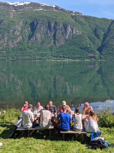 Sosial samling på benk i sommarvêr ved grøn fjord med fjell bak