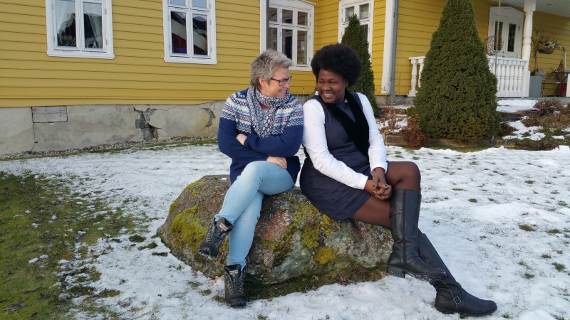 To kvinner sit på ein stein med snø rundt. Dei har beina i kors og ser på kvarandre med eit smil. Gult hus bak.